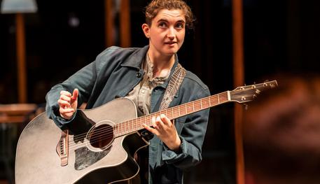 Kit Kenneth (Balthasar) in Much Ado About Nothing. Photo by Johan Persson. Kit wears a blue jacket and holds a black acoustic guitar, held over their shoulder with a strap decorated with cobwebs. They are about to begin a song.