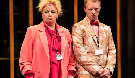 Caroline Parker (Dogberry) and Lee Farrell (Verges) in Much Ado About Nothing. Photo by Johan Persson. Caroline wears a vibrant pink suit and floral headband and Lee wears a floral pink suit and holds a bunch of flowers. They both look concerned.