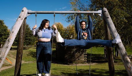 Two young people play on swings in a park on a sunny day, smiling and goofing around.