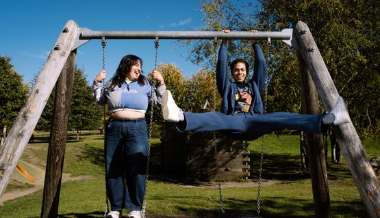 Two young people play on swings in a park on a sunny day, smiling and goofing around.