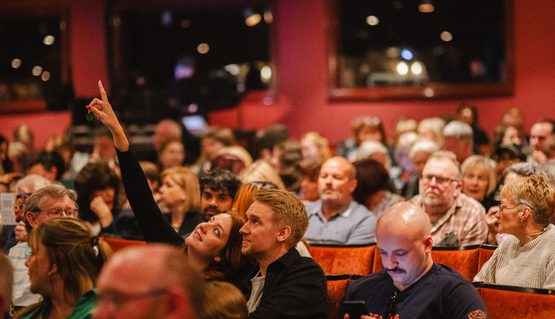 A couple smile and point upwards as they sit in orange seats in a theatre auditorium