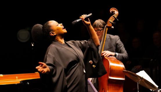 The celebration concert for Lord Bob Kerslake. Photo by Lucy Smith Jones. A woman wearing black sings passionately into a microphone. behind, A cellist plays/