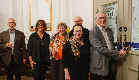 A group of people smile as a person at the front cuts a blue ribbon on the front of a pale green set of doors.