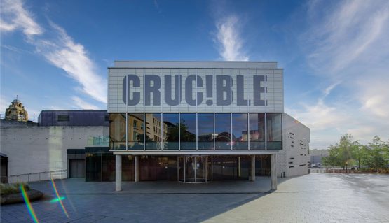 The front of the Crucible theatre: a brutalist building stands in the sunshine with blue sky behind it. There is a glass panelled room held up by concrete pillars, with a panel above which reads in capital letters: CRUC!BLE.