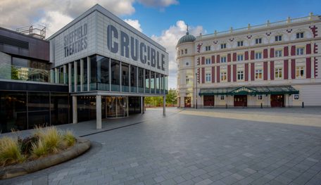 The Crucible and Lyceum theatres in the sunshine with blue sky behind. The Crucible is a brutalist building with a glass panelled room held up by concrete pillars and a panel above which reads in capital letters: CRUC!BLE. The Lyceum is a grand Victorian theatre, painted cream and burgundy. It has a rounded corner topped with a green lead turret with a statue of Mercury at the very peak.