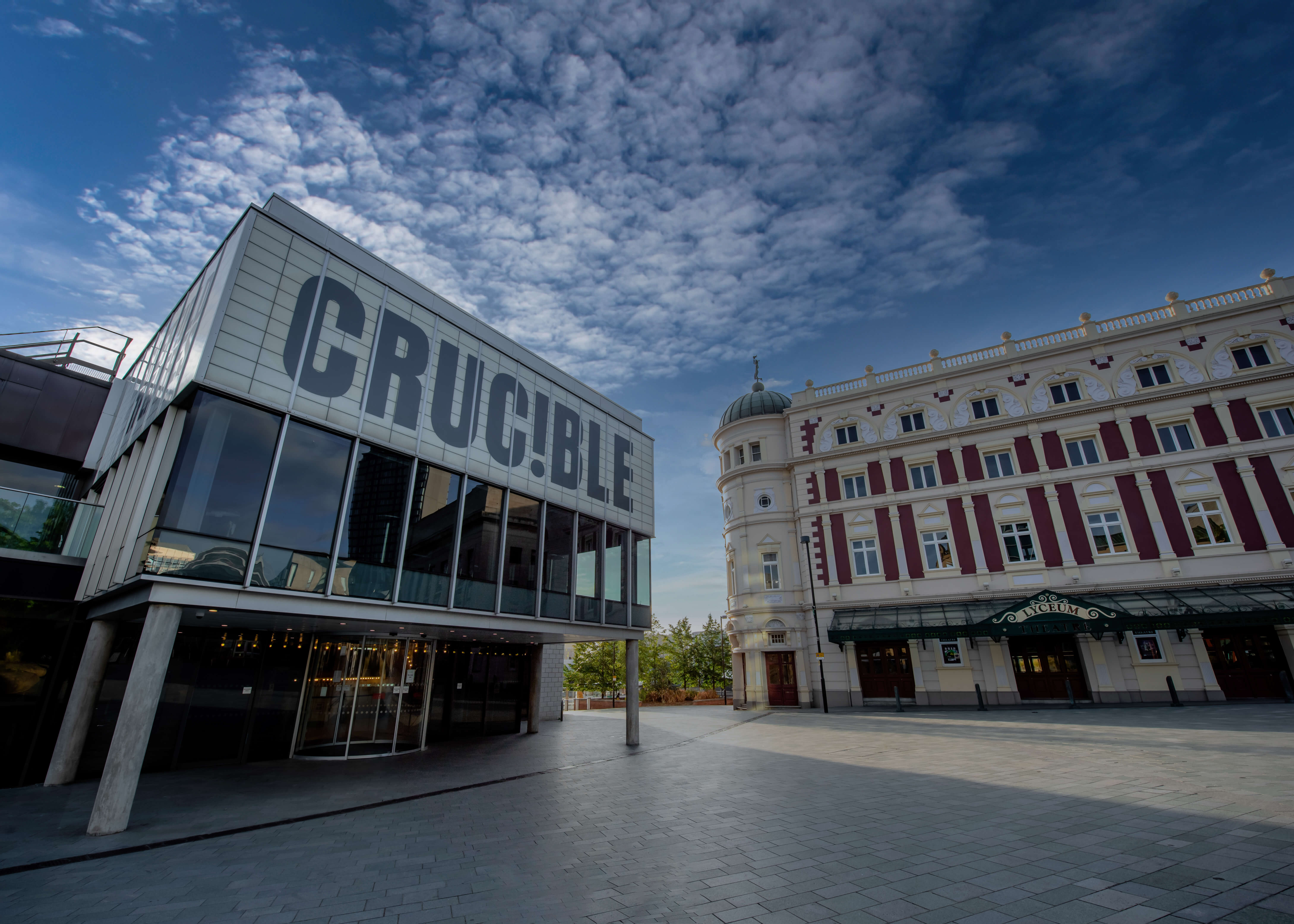 The Crucible and Lyceum theatres in the sunshine with blue sky behind. The Crucible is a brutalist building with a glass panelled room held up by concrete pillars and a panel above which reads in capital letters: CRUC!BLE. The Lyceum is a grand Victorian theatre, painted cream and burgundy. It has a rounded corner topped with a green lead turret with a statue of Mercury at the very peak.