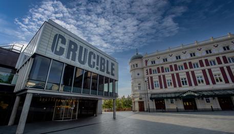 The Crucible and Lyceum theatres in the sunshine with blue sky behind. The Crucible is a brutalist building with a glass panelled room held up by concrete pillars and a panel above which reads in capital letters: CRUC!BLE. The Lyceum is a grand Victorian theatre, painted cream and burgundy. It has a rounded corner topped with a green lead turret with a statue of Mercury at the very peak.