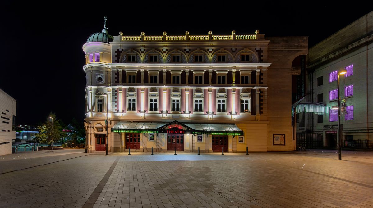 The Lyceum theatre lit up at night-time. The Lyceum is a grand Victorian theatre, painted cream and burgundy. It has a rounded corner topped with a green lead turret with a statue of Mercury at the very peak.