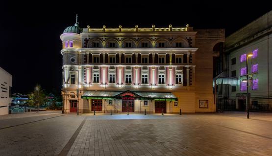 The Lyceum theatre lit up at night-time. The Lyceum is a grand Victorian theatre, painted cream and burgundy. It has a rounded corner topped with a green lead turret with a statue of Mercury at the very peak.