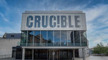 The front of the Crucible theatre: a brutalist building stands in the sunshine with blue sky behind it. There is a glass panelled room held up by concrete pillars, with a panel above which reads in capital letters: CRUC!BLE.