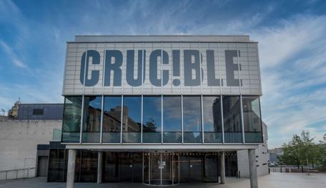 The front of the Crucible theatre: a brutalist building stands in the sunshine with blue sky behind it. There is a glass panelled room held up by concrete pillars, with a panel above which reads in capital letters: CRUC!BLE.