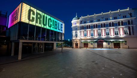 The Crucible and Lyceum theatres lit up at night-time. The Crucible is a brutalist building with a glass panelled room held up by concrete pillars and a panel above lit up in rainbow colours, which reads in capital letters: CRUC!BLE. The Lyceum is a grand Victorian theatre, painted cream and burgundy. It has a rounded corner topped with a green lead turret with a statue of Mercury at the very peak.