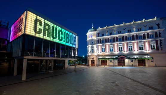 The Crucible and Lyceum theatres lit up at night-time. The Crucible is a brutalist building with a glass panelled room held up by concrete pillars and a panel above lit up in rainbow colours, which reads in capital letters: CRUC!BLE. The Lyceum is a grand Victorian theatre, painted cream and burgundy. It has a rounded corner topped with a green lead turret with a statue of Mercury at the very peak.