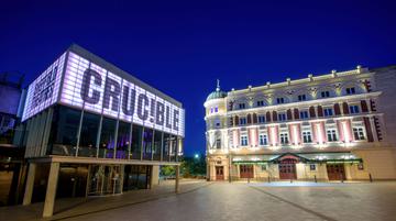 The Crucible and Lyceum theatres lit up at night-time. The Crucible is a brutalist building with a glass panelled room held up by concrete pillars and a panel above lit up in white, which reads in capital letters: CRUC!BLE. The Lyceum is a grand Victorian theatre, painted cream and burgundy. It has a rounded corner topped with a green lead turret with a statue of Mercury at the very peak.