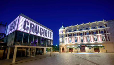 The Crucible and Lyceum theatres lit up at night-time. The Crucible is a brutalist building with a glass panelled room held up by concrete pillars and a panel above lit up in white, which reads in capital letters: CRUC!BLE. The Lyceum is a grand Victorian theatre, painted cream and burgundy. It has a rounded corner topped with a green lead turret with a statue of Mercury at the very peak.