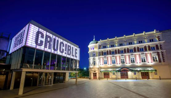 The Crucible and Lyceum theatres lit up at night-time. The Crucible is a brutalist building with a glass panelled room held up by concrete pillars and a panel above lit up in white, which reads in capital letters: CRUC!BLE. The Lyceum is a grand Victorian theatre, painted cream and burgundy. It has a rounded corner topped with a green lead turret with a statue of Mercury at the very peak.