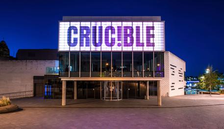 The front of the Crucible theatre: a brutalist building in the darkness, lit up in white light. There is a glass panelled room held up by concrete pillars, with a lit up panel above which reads in capital letters: CRUC!BLE.