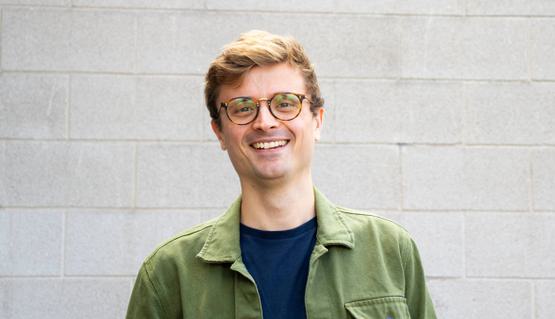 A man smiles to the camera. They are wearing glasses and a green shirt