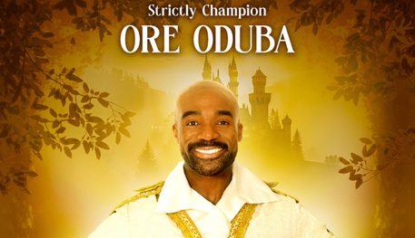 A person with a beard smiles and holds their hands on their hips, wearing a white and gold shirt. Text reads: STRICTLY CHAMPION ORE ODUBA