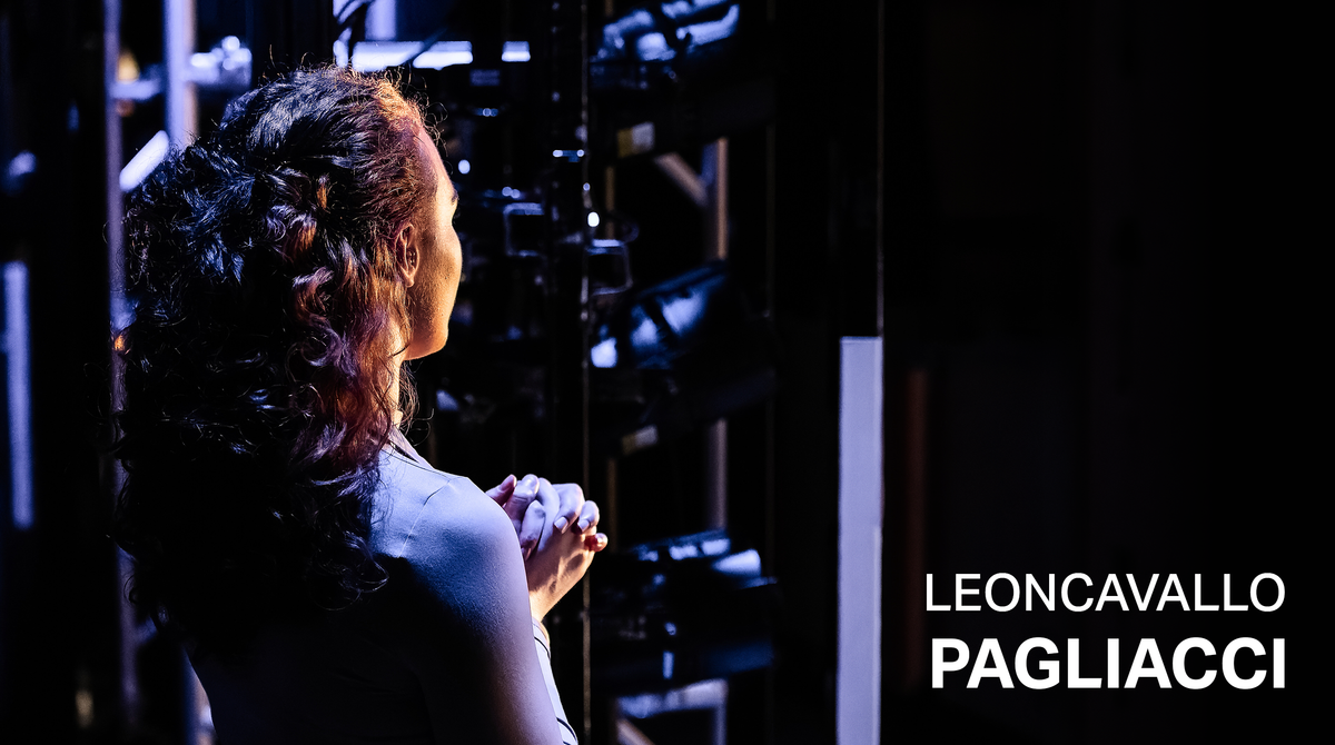Text reads: 'Leoncavallo Pagliacci'. A woman standing to the side of the stage. She has her back turned to the camera.
