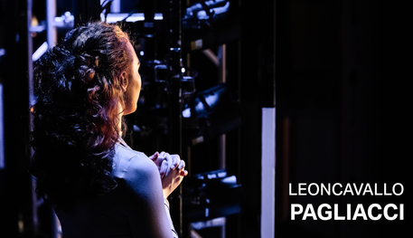 Text reads: 'Leoncavallo Pagliacci'. A woman standing to the side of the stage. She has her back turned to the camera.