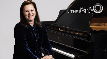 Kathryn Stott is sitting next to a grand piano. She is wearing a dark shirt and is smiling.