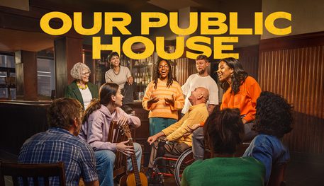 Text reads: 'Our Public House'. There are ten people of mixed ages in a pub. There is a woman in the centre who is talking and everyone is watching her and smiling. One woman is holding a guitar.