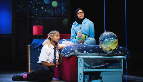 Jamie who is wearing schoool uniform and bright pink socks, sits on the floor leaning on Pritti's bed. Pritti sits on the bed. She wears a light blue jacket, a black headscarf and hold a Buzz Lightyear toy.