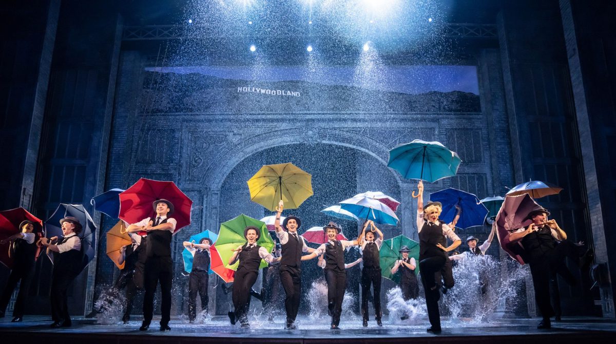 The cast of Singin' in the Rain are on stage holding umbrella's and dancing as water splashes up from the stage
