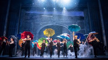 The cast of Singin' in the Rain are on stage holding umbrella's and dancing as water splashes up from the stage