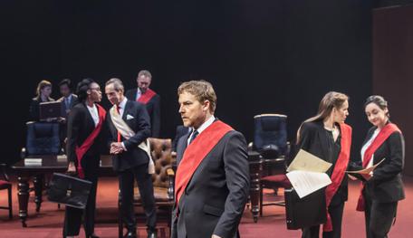 Sam West as Brutus wearing a suit and red sash. Behind him are other members of the senate. He looks thoughtful
