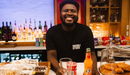 A person smiles behind a counter. Cakes can be seen on the counter and drinks and beer taps are behind them.