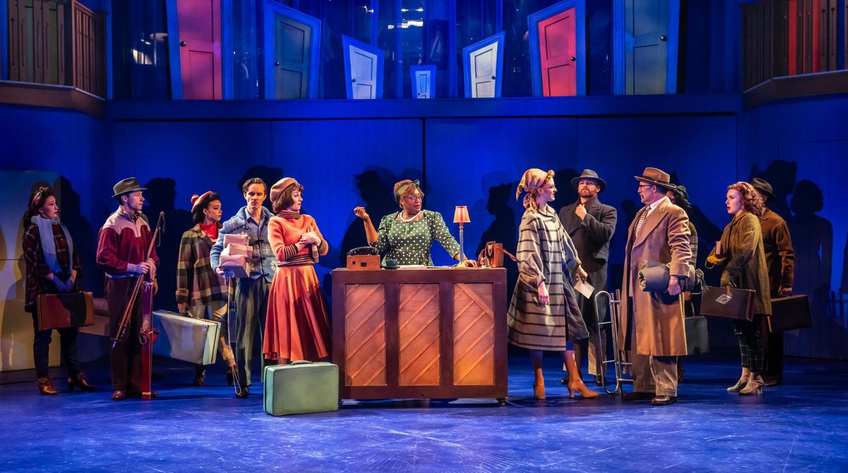 A production photo of the cast of White Christmas. A woman stands behind a desk in the middle of the image as ten people gather around here with suitcases looking annoyed.