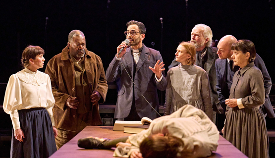 The company of The Crucible directed by Antony Lau. Left to Right: Laura Pyper (Ann Putnam/Sarah Good), Geoffrey Aymer (Giles Corey), Sid Sagar (Reverend Hale), Rose Shalloo (Abigail Williams), Andrew Macbean (Francis Nurse), Mark Weinman (Thomas Putnam), Alexandra Mathie (Rebecca Nurse), Honor Kneafsey (front, Betty Parris) in The Crucible. A young woman lies slumped on a table. At the end of the table, a group of people look confused and concerned. A man speaks into a microphone and reassures the group.