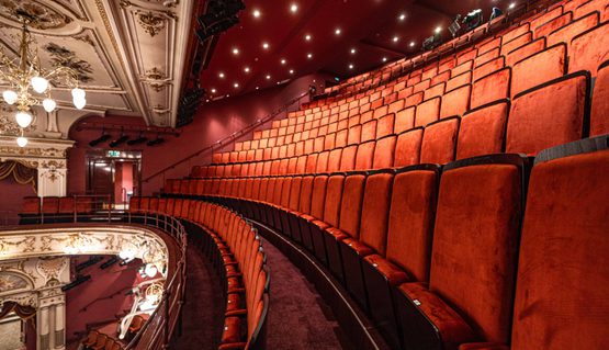 Velvet orange chairs in a Victorian theatre balcony. Light shine down from above.
