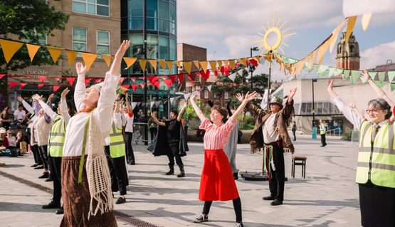 A group of performers stand with their arms up in a sunny outdoor square. Yellow bunting festoons above them.