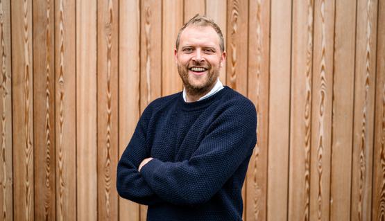 Tom Bird smiles to the camera, standing with arms folded in front of a wood panelled wall, he is wearing a navy blue jumper
