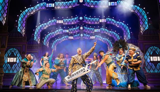 The cast of Aladdin on stage dancing as the musical director stands in front with a keyboard singing