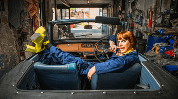 An image of a man in drag, wearing large yellow heels and a ginger wing, sitting in the back of a car with his legs up. The car is parked in a garage. He is biting his finger and looking at the camera.