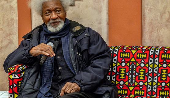 An elderly person with grey hair and a grey beard sits on an African patterned sofa and smiles, holding a microphone