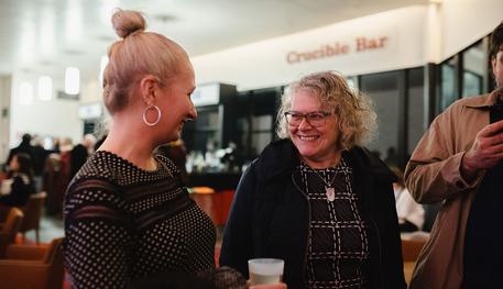 Two women talking and smiling outside of the Crucible Bar. Both women are older and have blonde hair. The woman on the left is wearing a black and white dress and the woman on the right is wearing a dark blue coat and a black and white square dress.