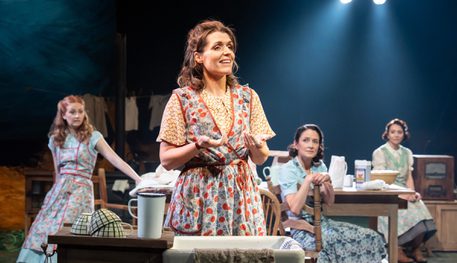 A person wearing a floral apron holds their hands over a sink which are covered in flour. They smile as they speak, and three others behind watch them, smiling.