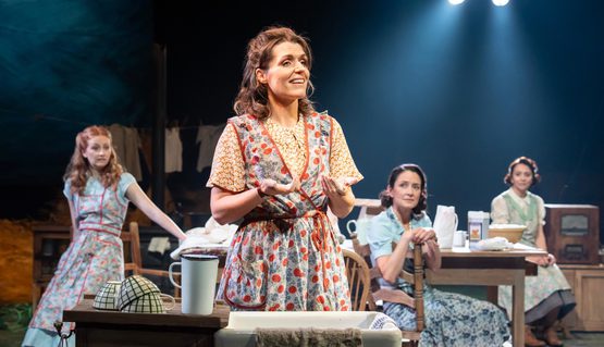 A person wearing a floral apron holds their hands over a sink which are covered in flour. They smile as they speak, and three others behind watch them, smiling.