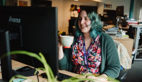 A person holds a mug and smiles as they type, looking at a computer screen.