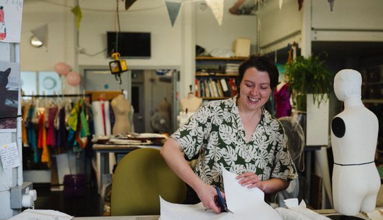 A person smiles as they cut material on a table. Behind them, costumes and mannequins can be seen.